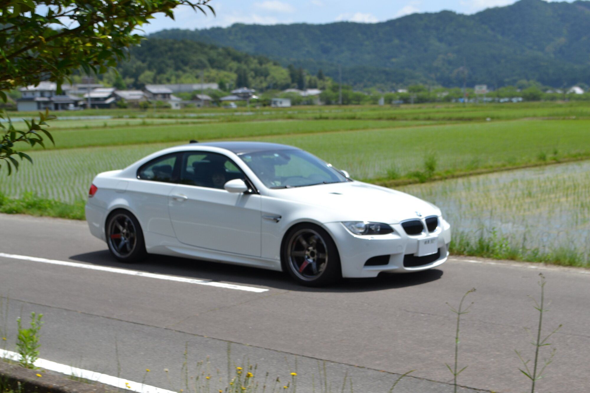 White BMW M3 E92 coupe driving past rice fields, front three-quarter view
