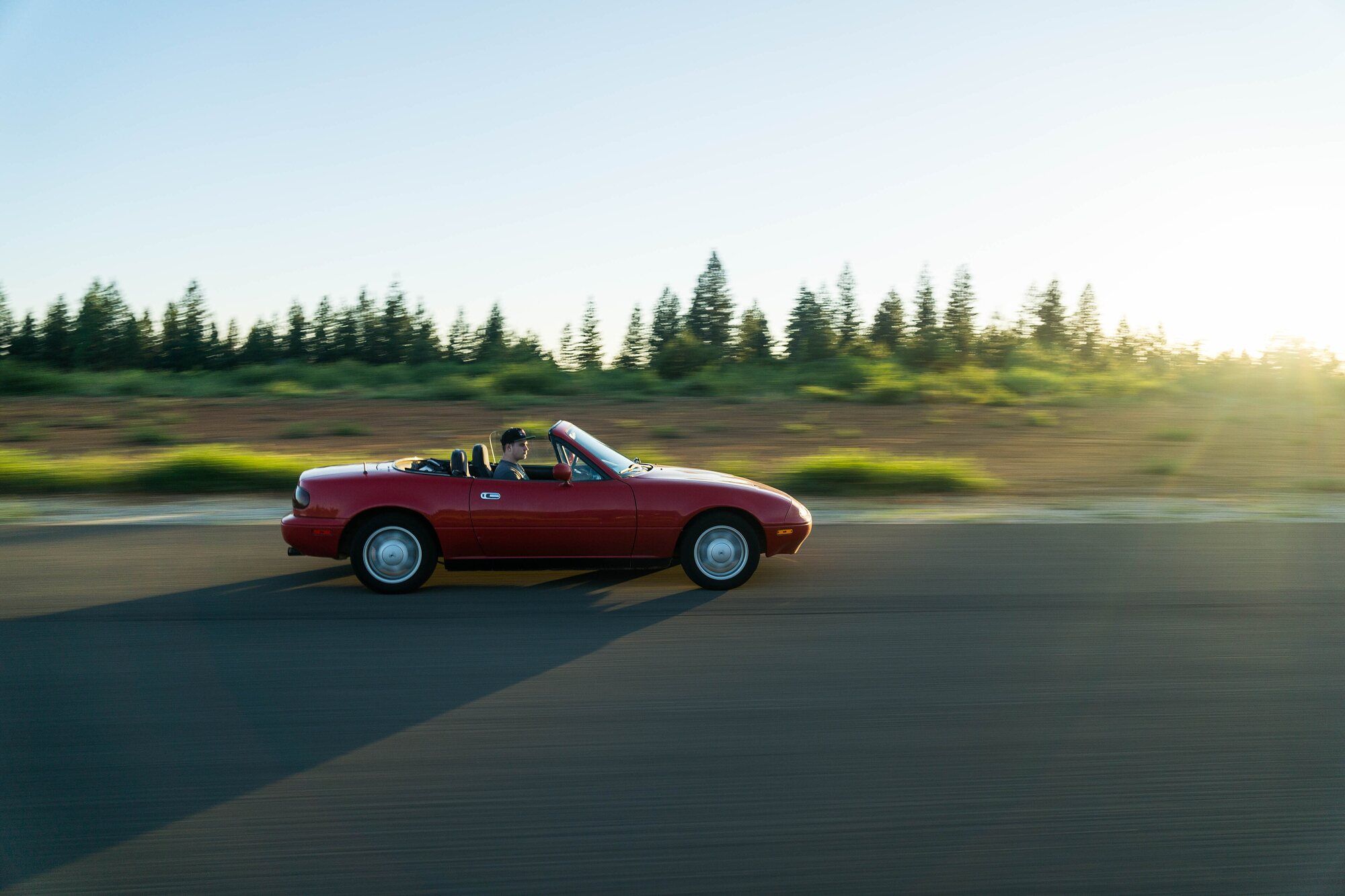 Red NA Mazda Miata driving on an empty two-lane road, seen from the side, top down